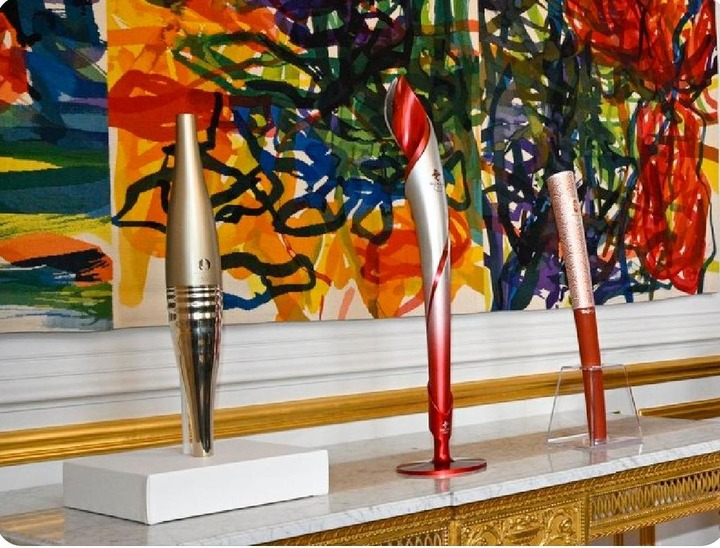 Three Olympic torches stand side by side on an off-white marble table in the brightly lit Portrait Room of Elysee Palace.